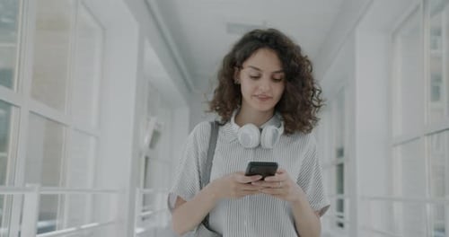 Dolly Shot of Happy Young Lady Student Walking in University Hall Using Smartphone Texting