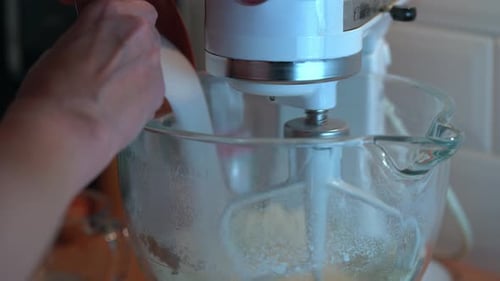 Woman adds sugar ingredient to a moving electric stand mixer with flour in glass bowl.
