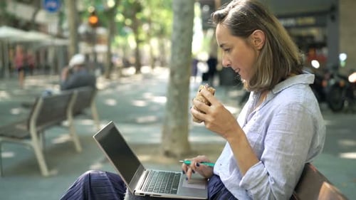 Junge Geschäftsfrau genießt Mittagessen mit Laptop in der Stadt auf einer sonnigen Bank