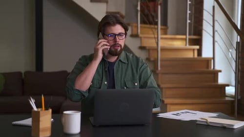 Young Bearded Businessman Talking on Phone and Sitting at Table with Laptop at Home or Office Spbas