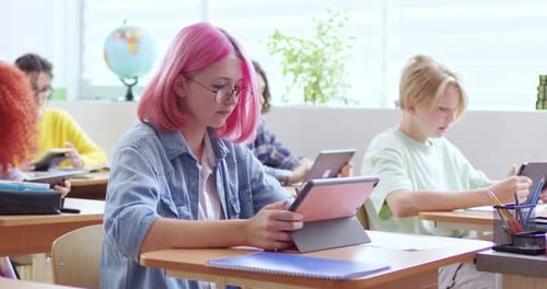 Teenage Caucasian Girl Student in Glasses with Pink Hair Sitting at Desk in Classroom at Lesson and