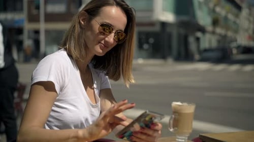 Young Woman with Coffee Texting on Smartphone Sitting in Cafe in City 30s
