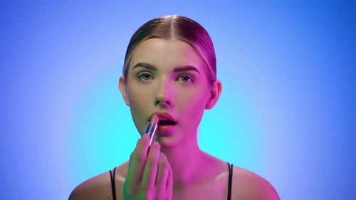 Caucasian young blonde girl paints her lips looking at camera, close up shot in beauty studio