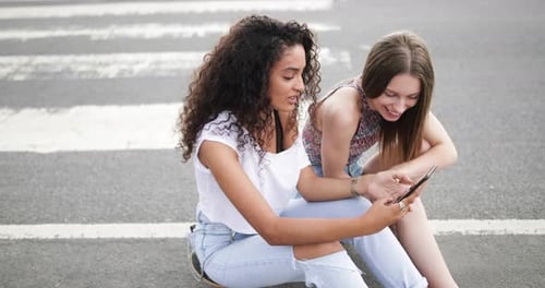 Two Young Women Sitting on Street Looking at Phone