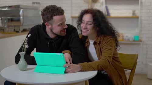Joyful Boyfriend and Girlfriend Talking Sitting at Table in Cafe Surfing Internet on Digital Tablet