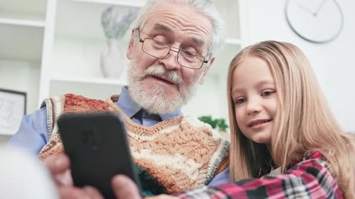 Grandfather and Granddaughter Look at Smartphone Together Indoors