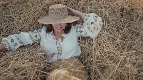 Pretty Woman in Straw Hat and Embroidered Blouse Smiling Lying on Hay in Countryside at Sunset