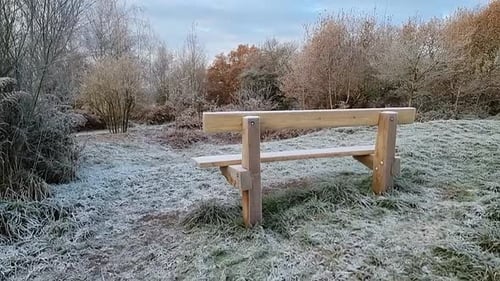 Empty wooden park bench on frosty ice coated grassy hillside overlooking autumn woodland foliage