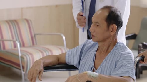 Medical team helping an elderly Asian man in a wheelchair moving through hospital hallway