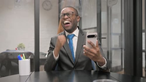 African Businessman Excited for Win on Phone at Workspace