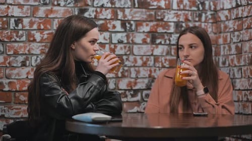 Two Brownhaired Girls Talk in a Cafe Over Glasses of Juice