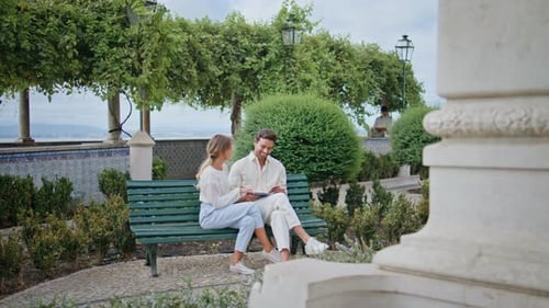 Recién casados sonrientes leyendo un libro en Greenery Square Bench Lovers Talking Together