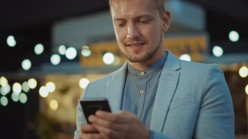 Handsome Young Concentrated Man is Using a Smartphone while Standing Outdoors in the Evening. He's