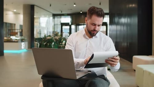 Focused Young Businessman Using Laptop and Analyzing Graph Documents Reports in Modern Office Lobby