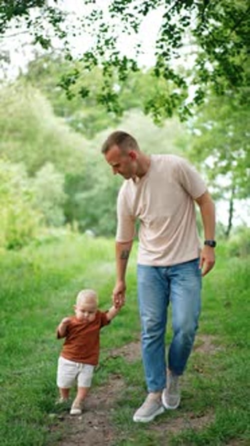 Father holds his little son by the hand walking by the park. Teaching a child to walk outdoors.