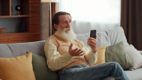 Vídeo de un hombre mayor barbudo chateando con amigos o familiares discutiendo noticias del mundo sentado en el sofá en