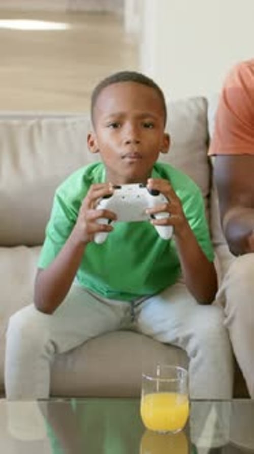 Vertikales Video: Spiel startet, Kind drückt Knöpfe auf dem Pad auf der Couch, trägt grünes T-Shirt, Saft