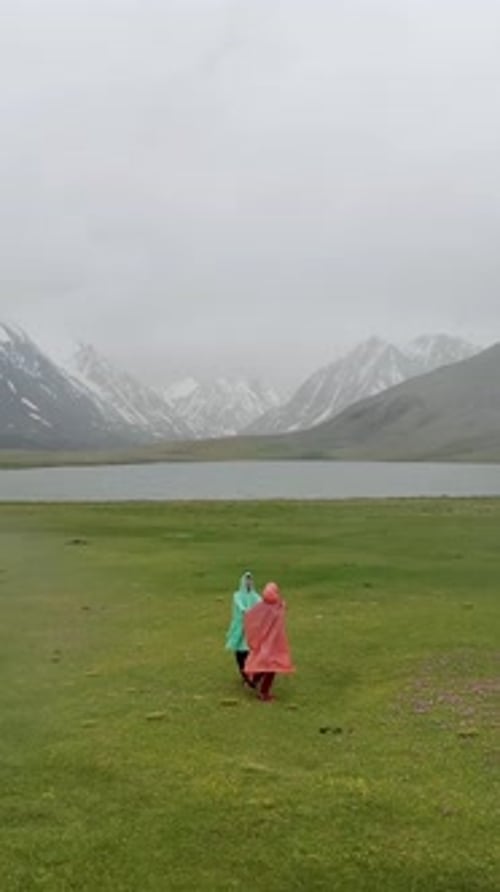 Couple of Tourists Holding Hands Standing in Grass in Rain on Background of Lake in Mountains Young