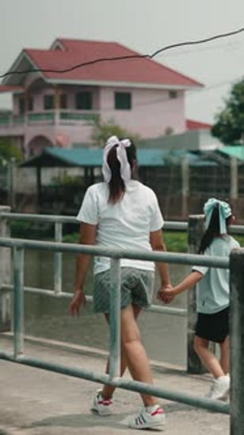 Young Woman and Child Walking Hand in Hand Along a Concrete Bridge By a Calm Riverside with Colorful