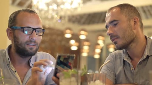 Two happy male friends chatting over smartphone in a cafe at night