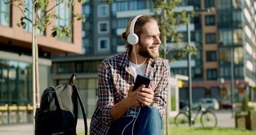 Cheerful Happy Young Caucasian Handsome Stylish Man in Headphones Sitting on Bench at Street and