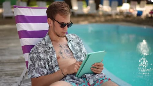 Slow Motion A Guy in Sunglasses Looks and Scrolls on the Tablet Screen While Sitting on a Beach