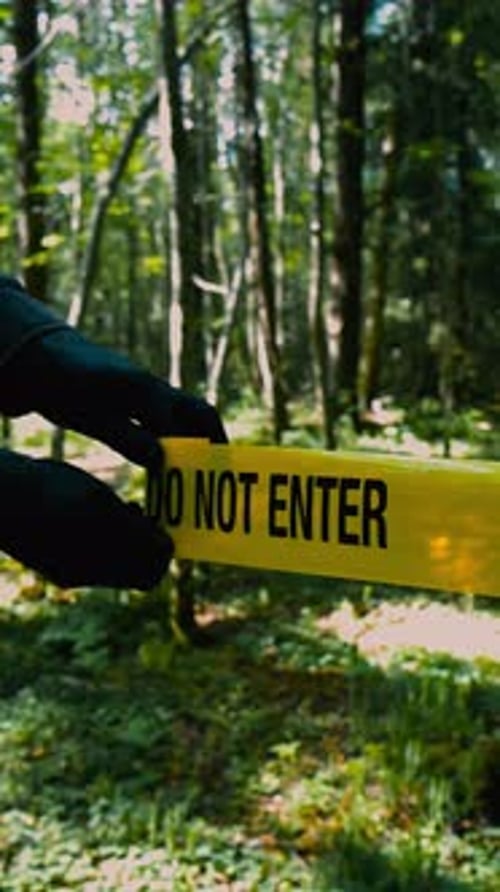 Detective with black gloves unrolling a Crime scene tape in front of a wooded forest area, sunny day