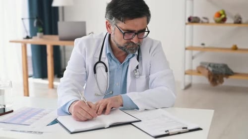 Writing information into notepad. Mature male doctor in coat is in the clinic room