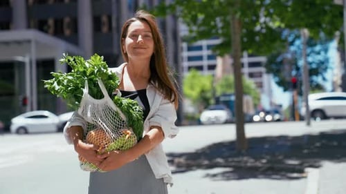 Una mujer sostiene productos frescos en una bolsa reutilizable mientras está de pie en una calle soleada del centro de la ciudad