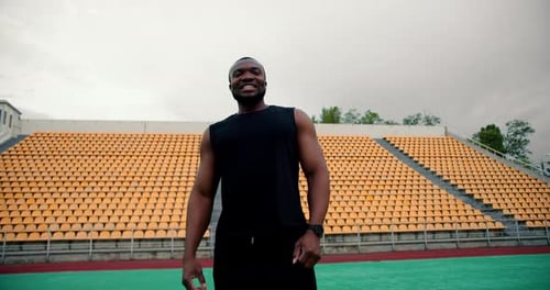 A Black Man in a Black Sports Uniform Poses and Uses a Battle Cry in Front of a Stadium with Yellow
