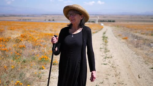 A happy old woman with gray hair walking on a hot dirt road in a summer field of flowers with a walk