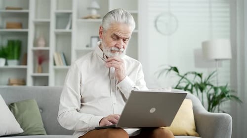 Serious thoughtfu elderly senior businessman working on laptop sitting on sofa in living room