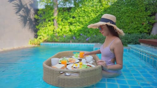 A young woman tourist has her own personal breakfast on a floating table in a private swimming pool.
