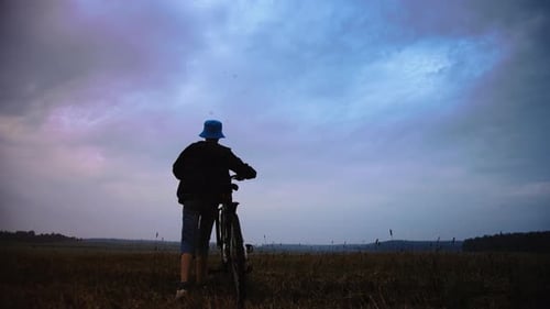Teen Boy With A Bicycle Walking In A Field In Rainy Summer Weather Goes Into the Distance Cinematic