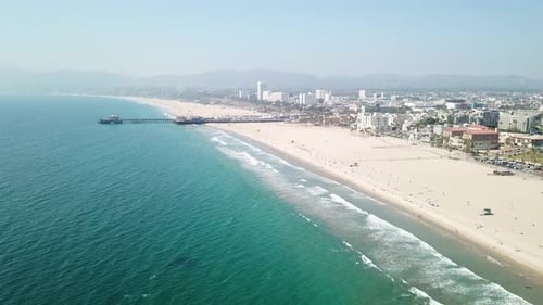 Sunny sandy Los Angeles beach skyline aerial rising towards pier above turquoise Pacific ocean