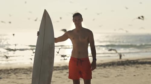Surfer Holding Surfboard on Beach at Sunset with Seagulls