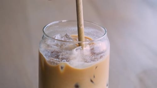 Close Up of Man Mixing Iced Latte in a Glass with a Straw on Cafe Table Cold Summer Refreshment