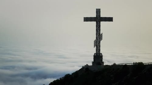 Towering Christian Cross Pierces the Sky Atop a Mountain Clouds Swirling Beneath It in a Timelapse