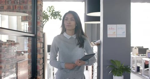 Biracial casual businesswoman walking through office with tablet, slow motion