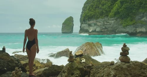 Seascape with contemplative woman. Adult girl in black swimsuit stands among rocks, sea behind.