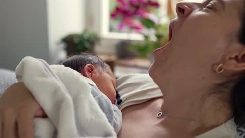 Mother yawning while holding her newborn baby close, illustrating the exhaustion and tender moments