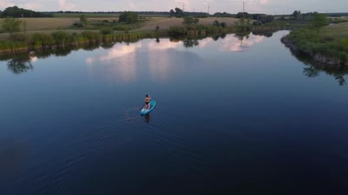 Drone View of a Young Woman in a Black Bikini Navigating a SUP Board on a Quiet Lake