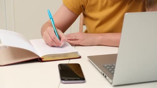 Woman Writes in Notebook at Desk With Laptop
