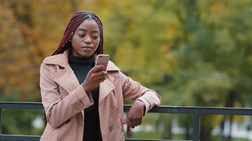 Young Serious Female Standing in Autumn Park Holding Mobile Phone Browsing Email Remotely