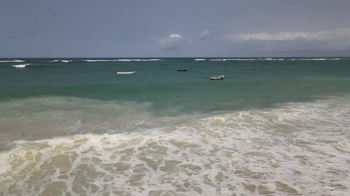 Schaumige Meereswellen rollen auf einen Sandstrand mit kleinen Fischerbooten, die im türkisfarbenen Wasser schwimmen