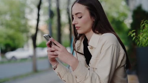 Young Woman Focused on Smartphone Outdoors in Park with Trees in the Background