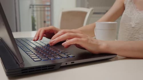 Close up shot of womans hands working on laptop and having a cup of tea nearby.