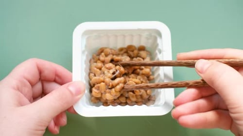Female hand holding chopsticks with natto beans on green background, top view.