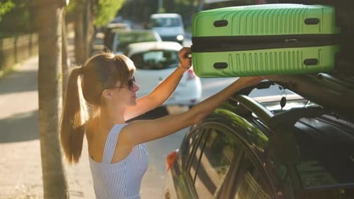 Young Woman Driver Putting Green Suitcase Inside Car Roof Rack on City Street