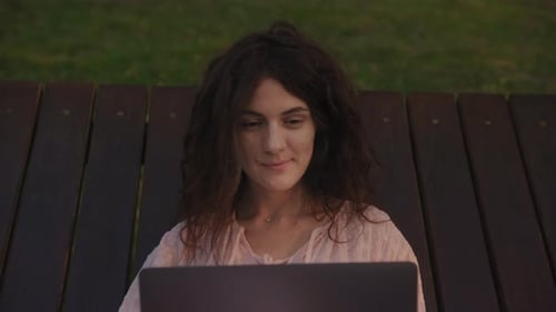 Handheld Close Up of a Curly Caucasian Woman Working on Her Laptop Computer on a Park Bench